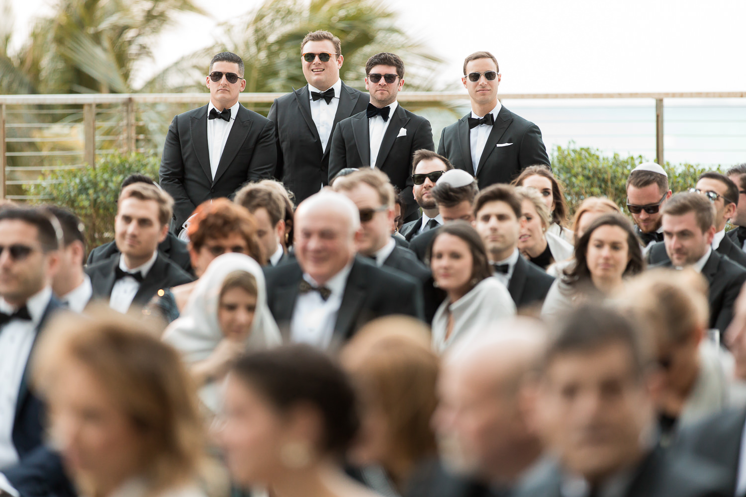 Groomsmen in black tuxedos watching a wedding ceremony.