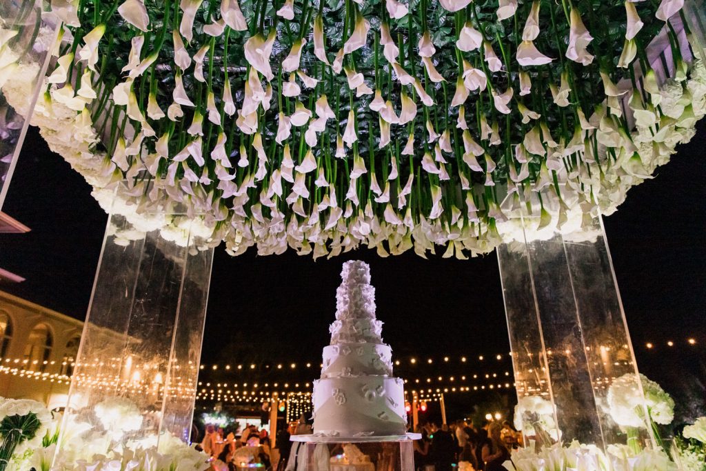 An all white wedding cake with flowers underneath a chuppah adorned with tulips.
