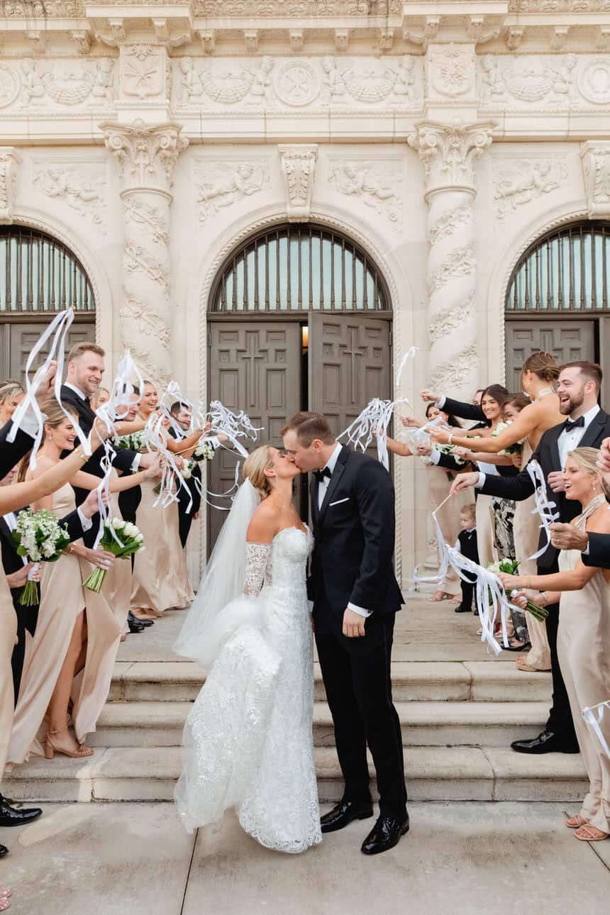 Bride and groom kiss on steps surrounded by friends celebrating with confetti and sparklers.