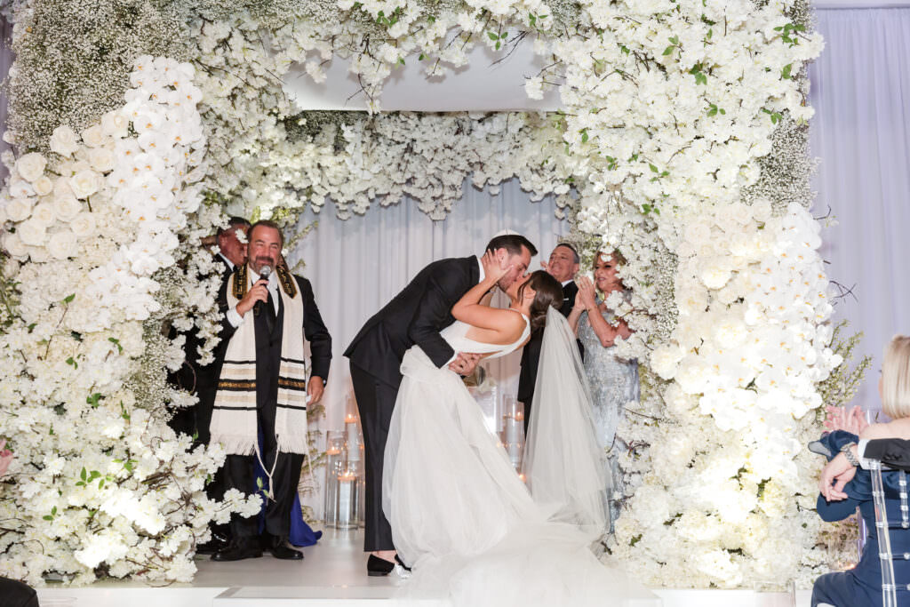 Bride and groom standing beneath a floral chuppah during outdoor wedding ceremony