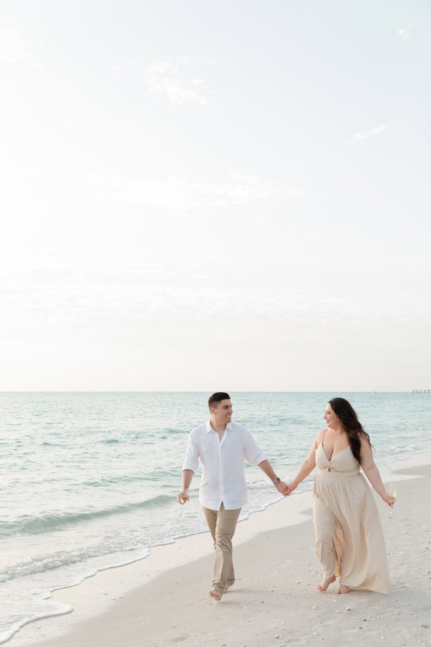 Bride and groom holding hands walking along the beach at sunset, smiling and enjoying the moment.