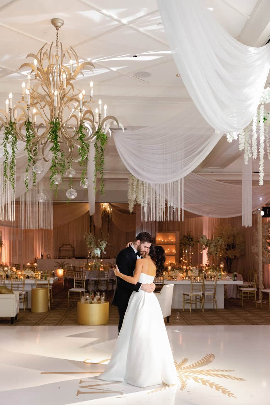 Bride and groom sharing a dance under elegant chandeliers and draped white fabric in a luxurious reception hall.