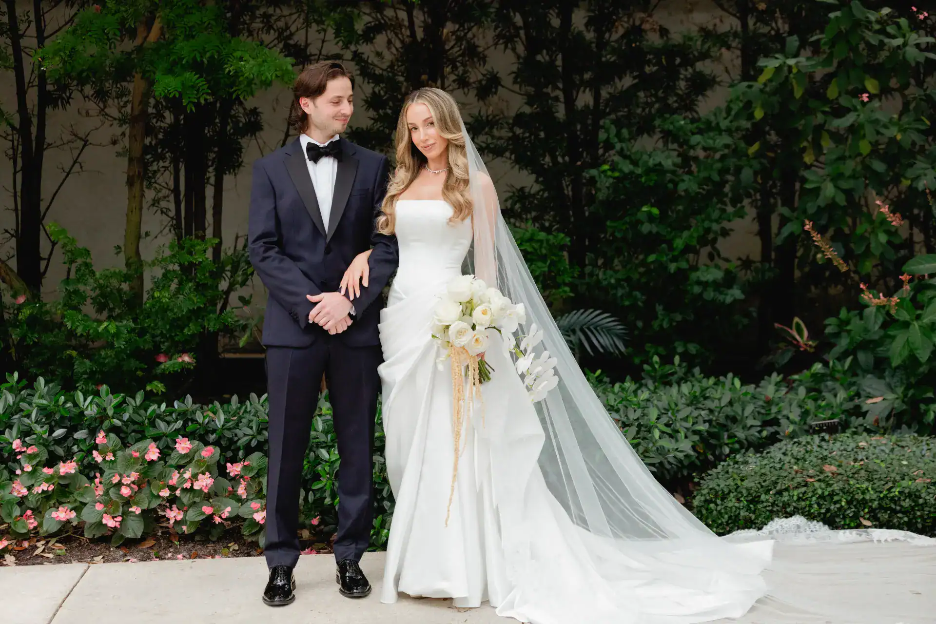 Bride and groom holding hands, smiling, standing outdoors with lush greenery and pink flowers.