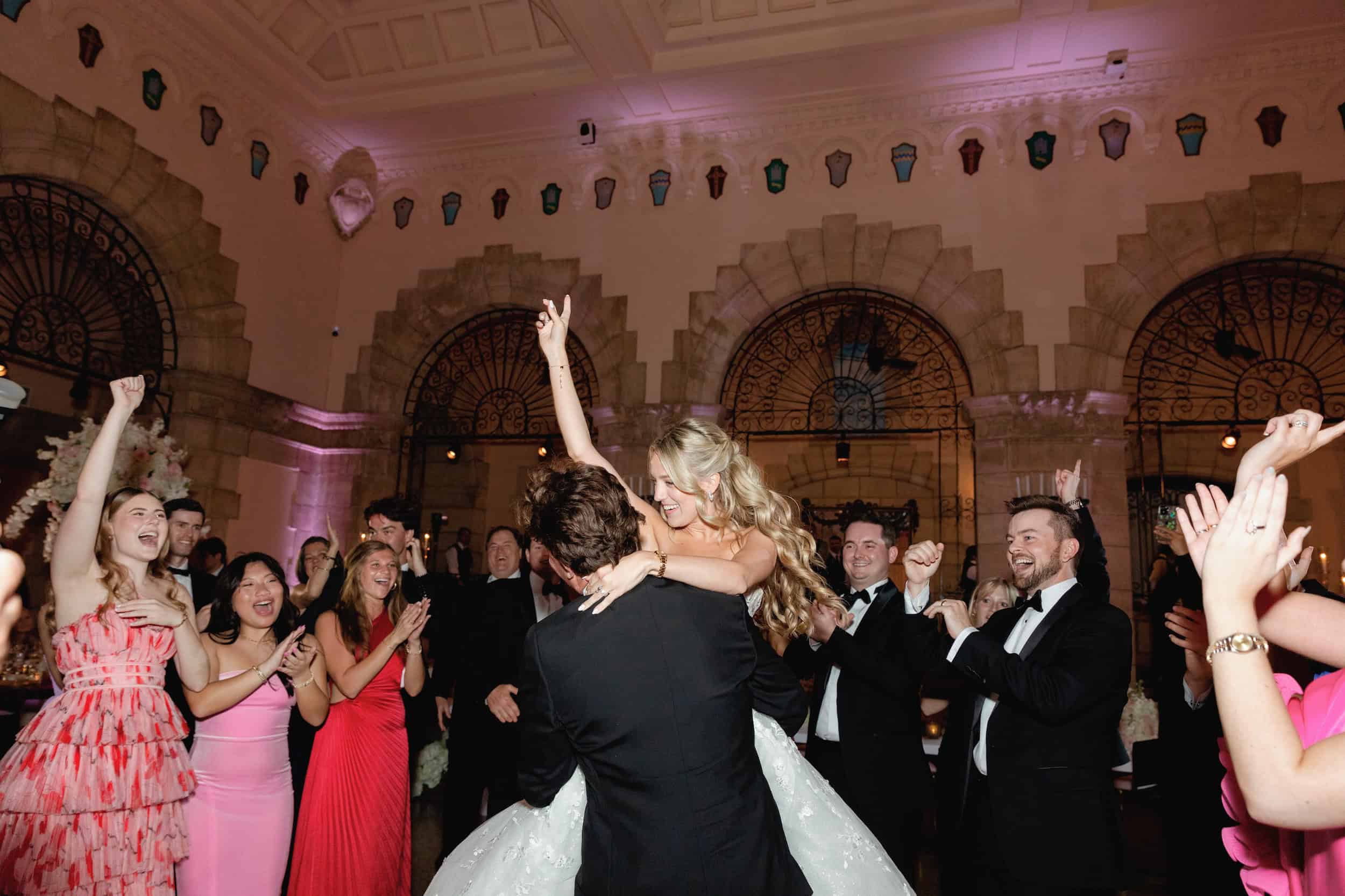 Bride and groom dancing surrounded by friends at a wedding reception with elegant architecture and warm lighting.