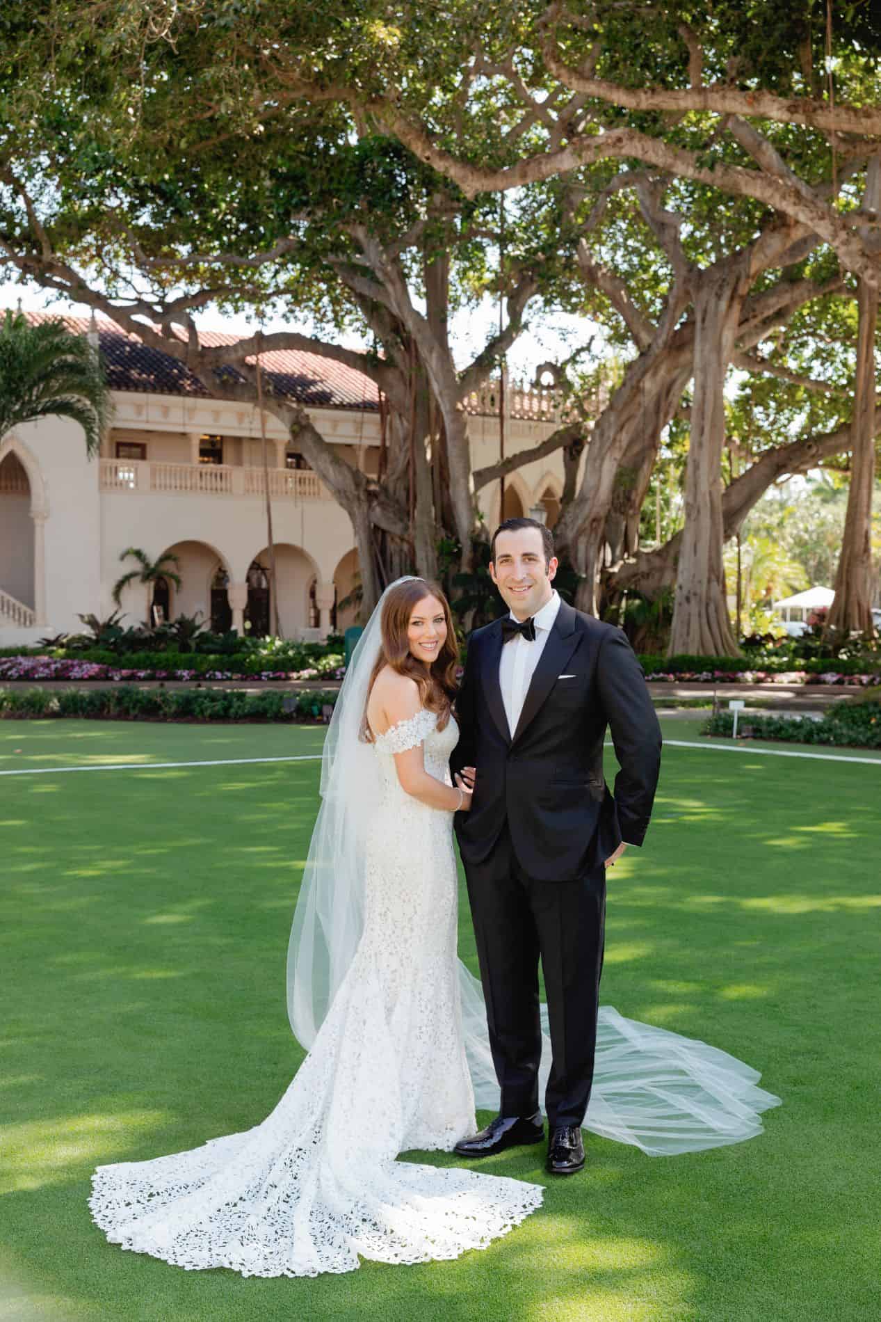 Bride and groom standing under a large tree, smiling, on a sunny day.