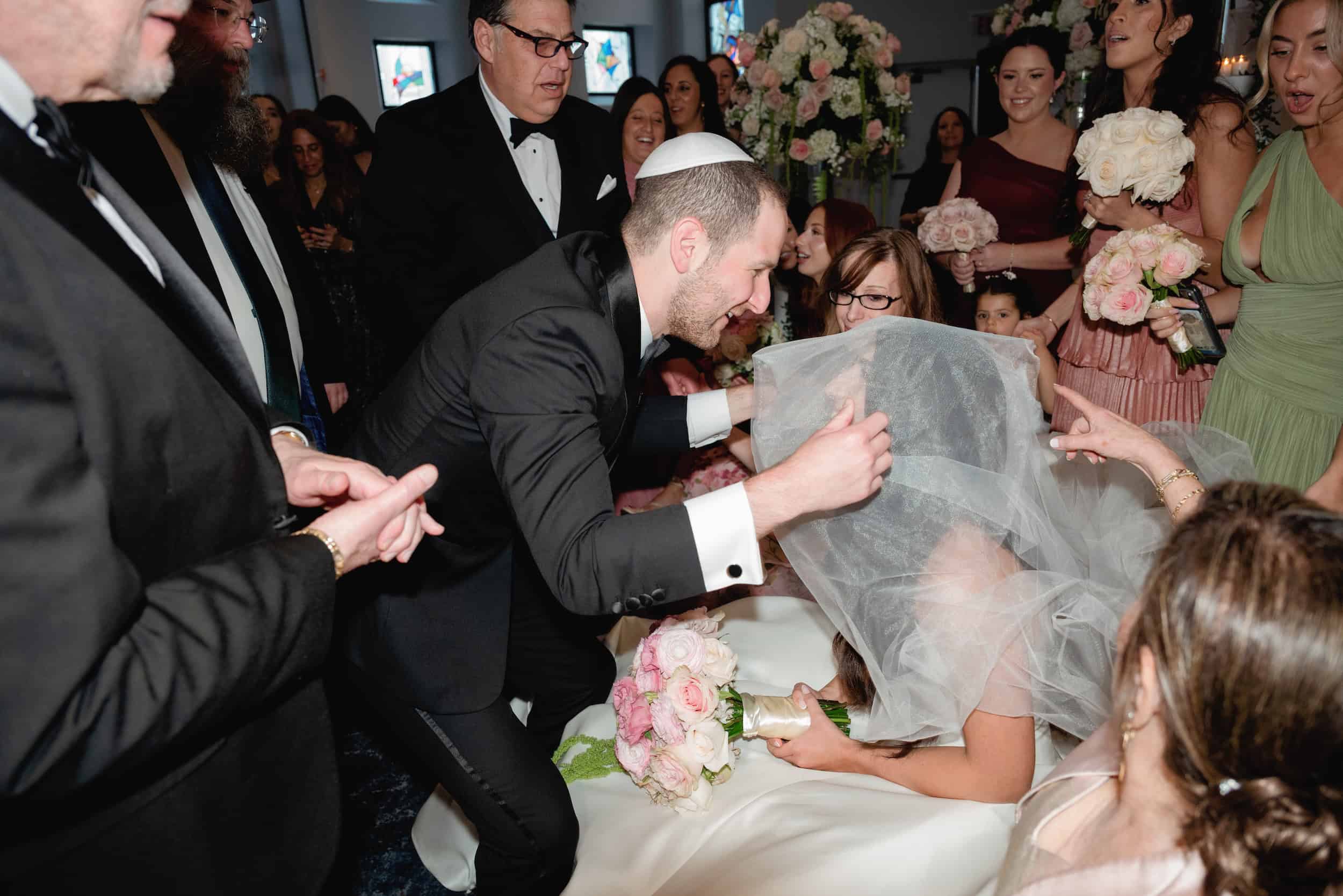 Groom in a tuxedo leans over to kiss the bride, who is seated, during a joyful moment at a wedding reception.