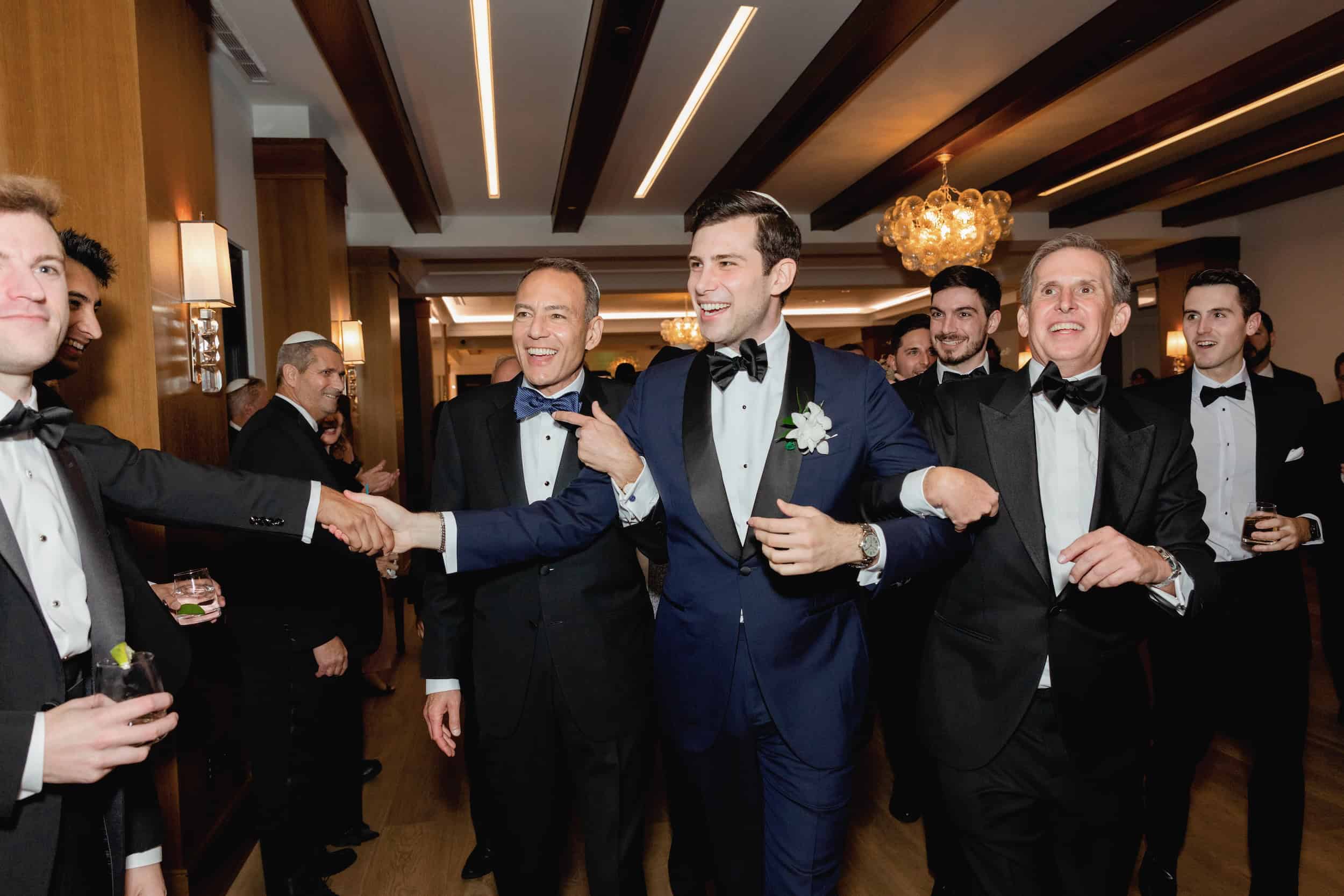 Groom in a navy tuxedo and bow tie greets guests at a wedding reception, smiling and shaking hands in a warmly lit venue.
