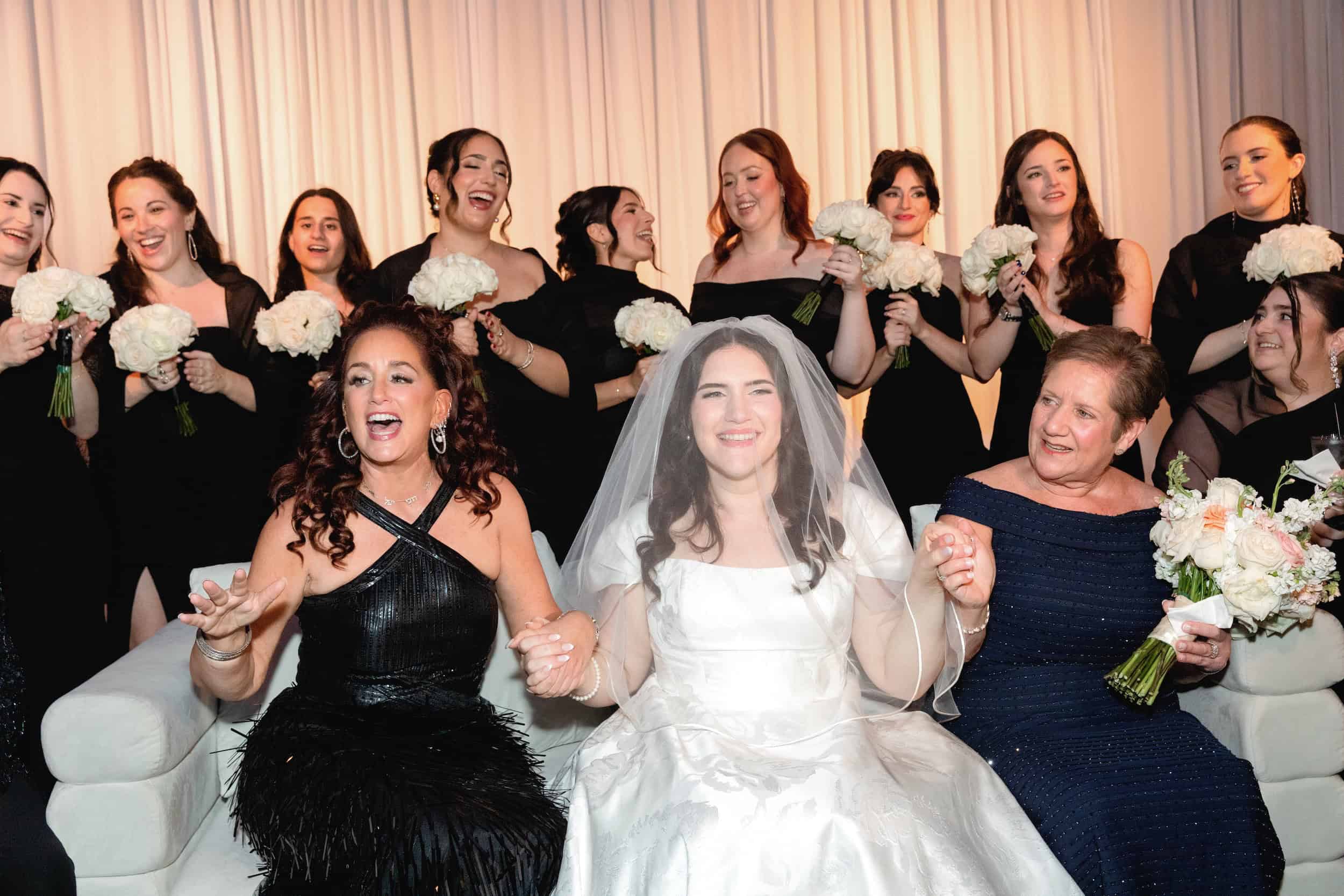 Bride in white gown smiling, surrounded by women holding bouquets, celebrating at a wedding reception.