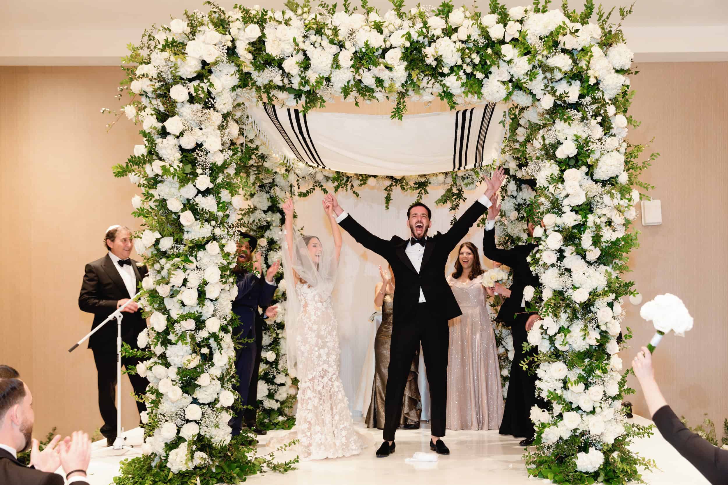 Bride and groom celebrating under a floral arch with friends and family at a wedding reception.