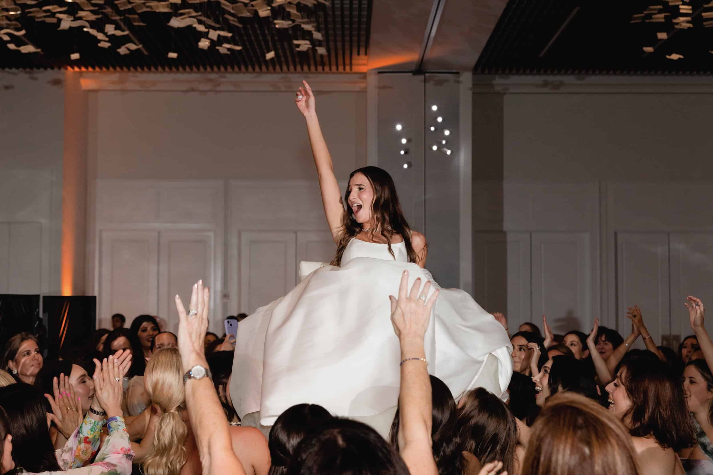Bride in white dress being lifted by guests at her wedding reception, joyful and celebratory atmosphere.