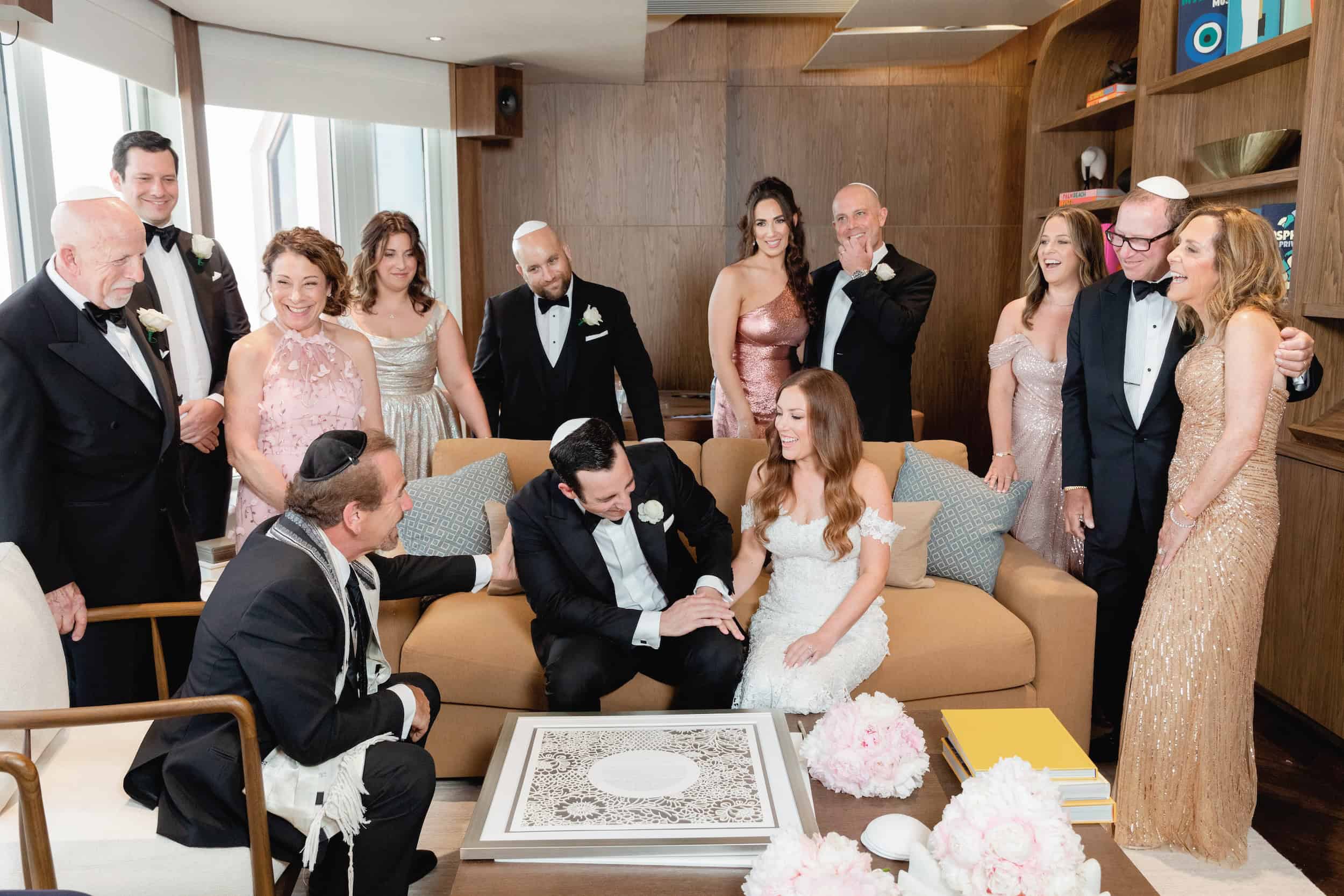 Bride and groom seated on a sofa, surrounded by family and friends in a modern living room, celebrating their wedding.
