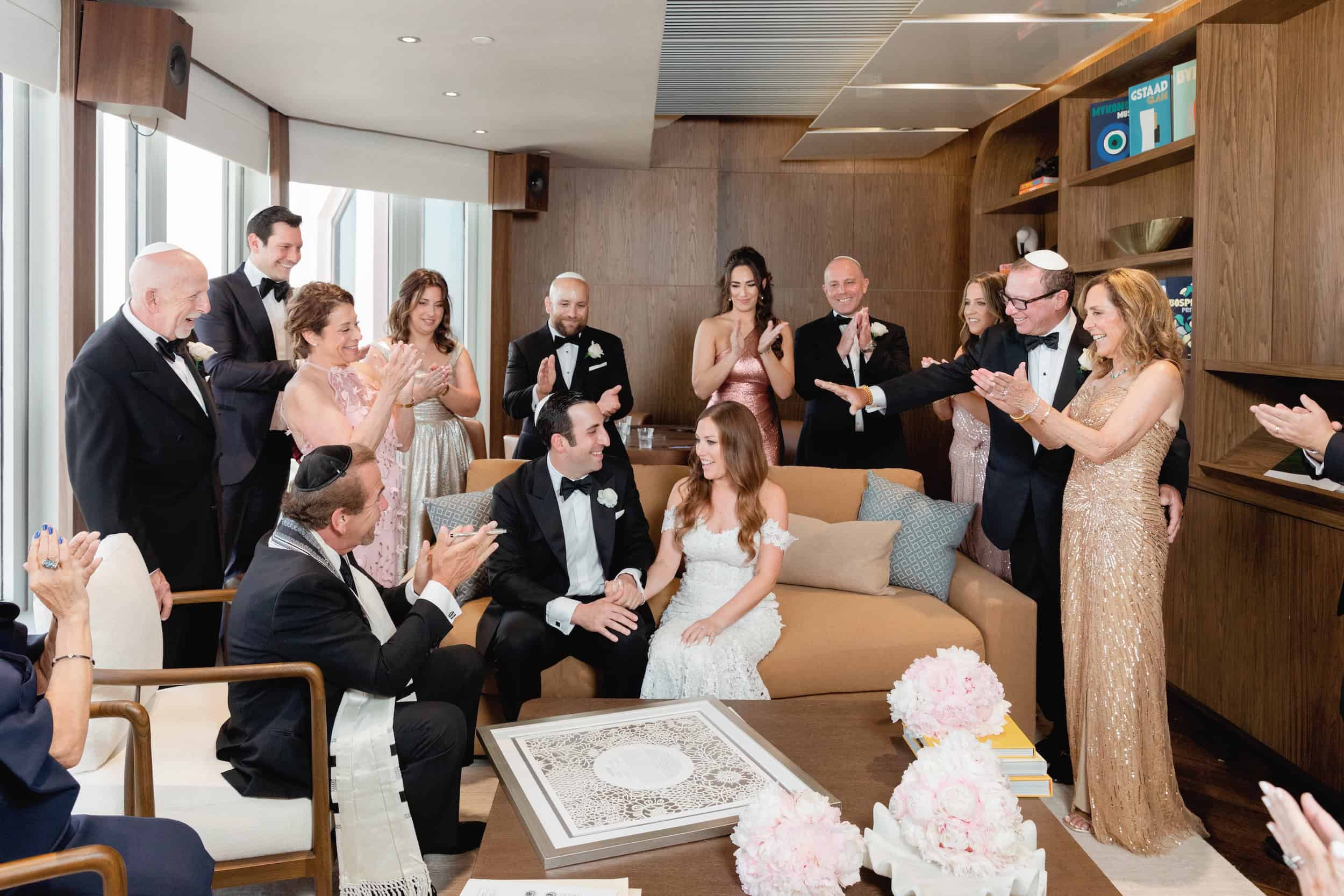 Bride and groom seated with family and friends in a modern living room, celebrating their wedding with applause and smiles.