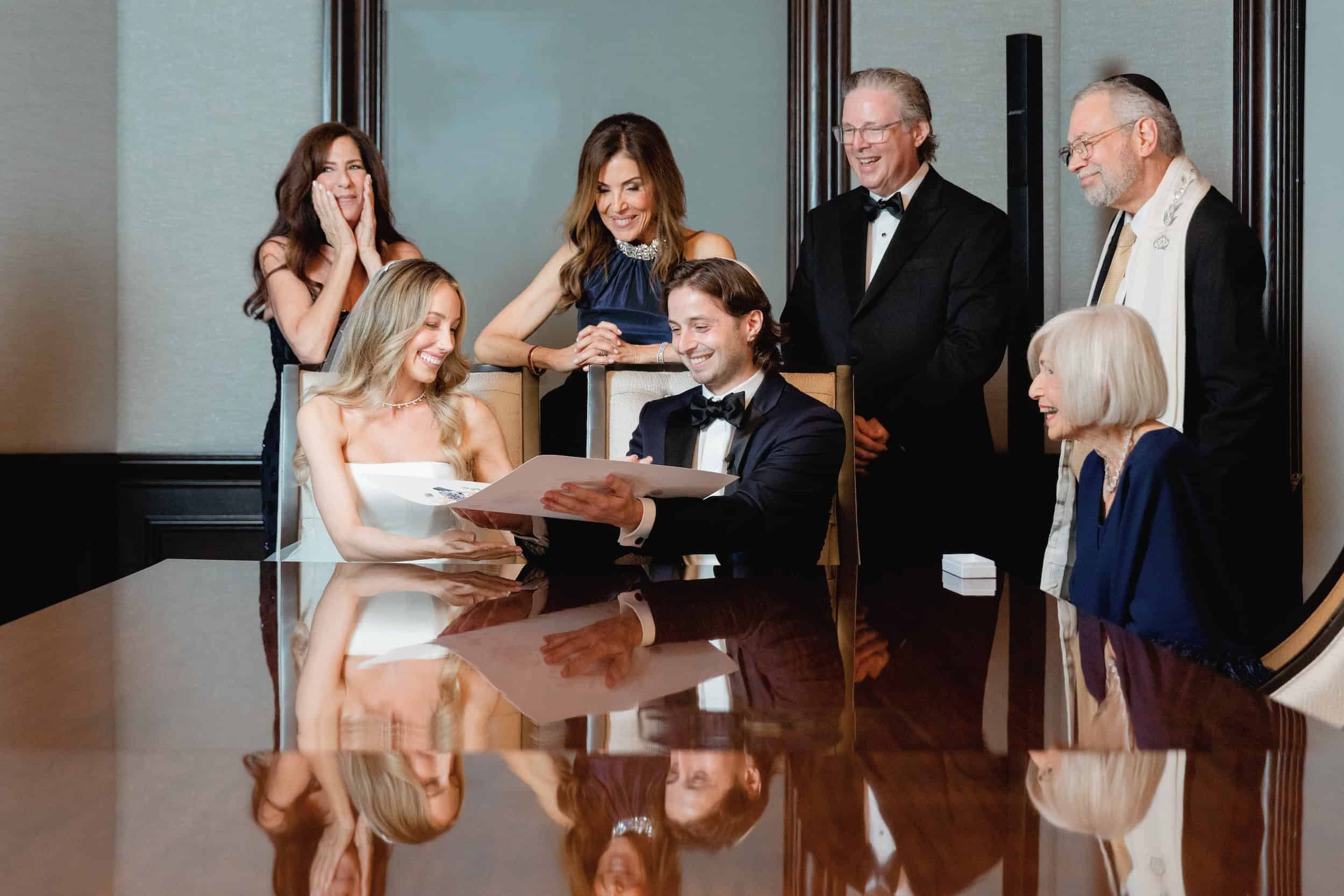 Bride and groom reading their wedding vows at a formal reception, surrounded by family and friends, with joyful expressions.