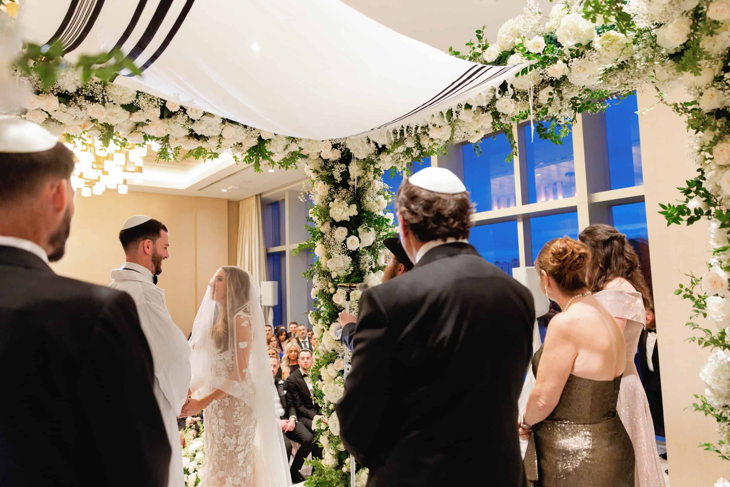 Bride and groom exchanging vows under floral arch with guests watching in a bright, elegant venue.