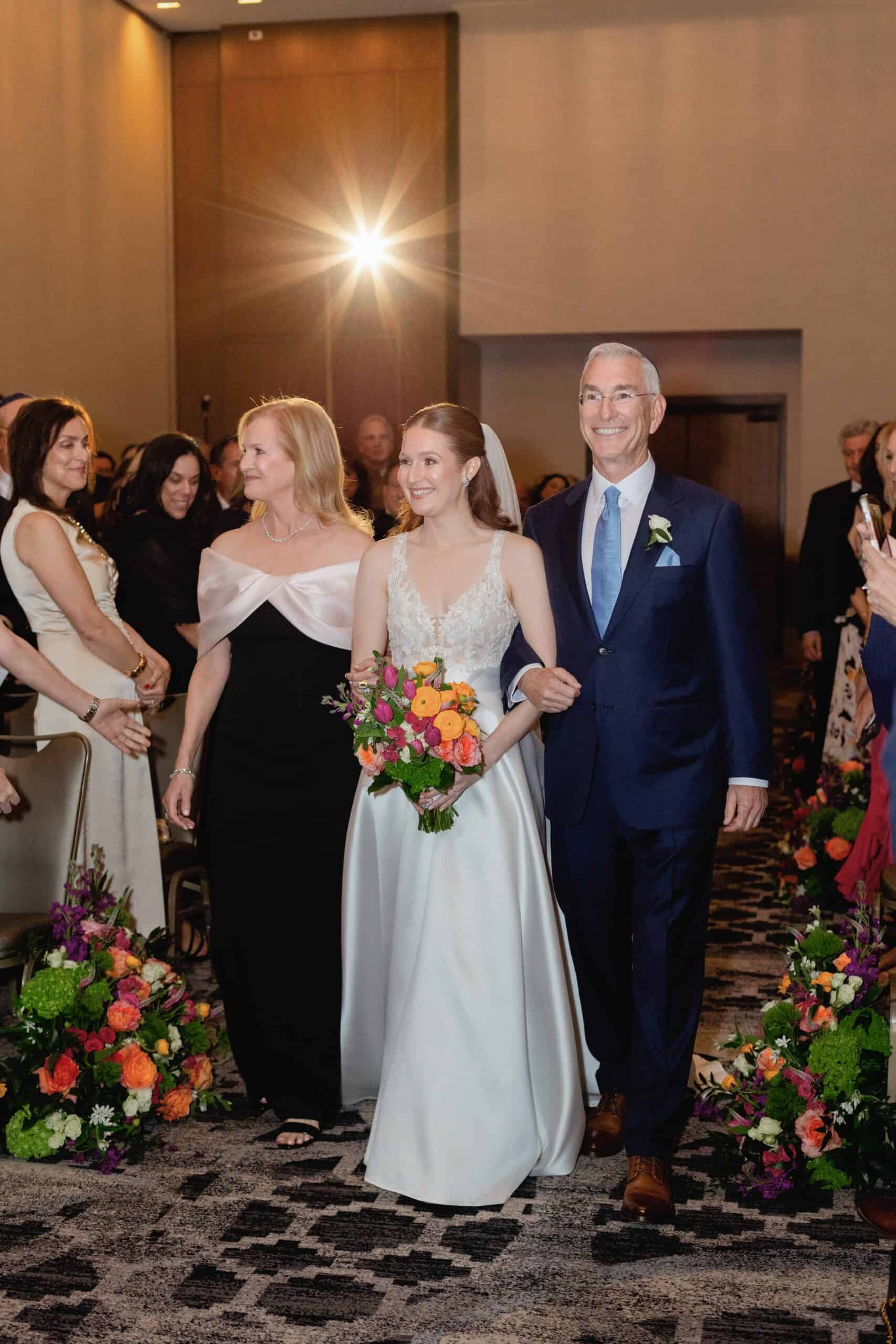 Bride and groom walking down the aisle, smiling, with guests on either side, floral arrangements, warm indoor lighting.