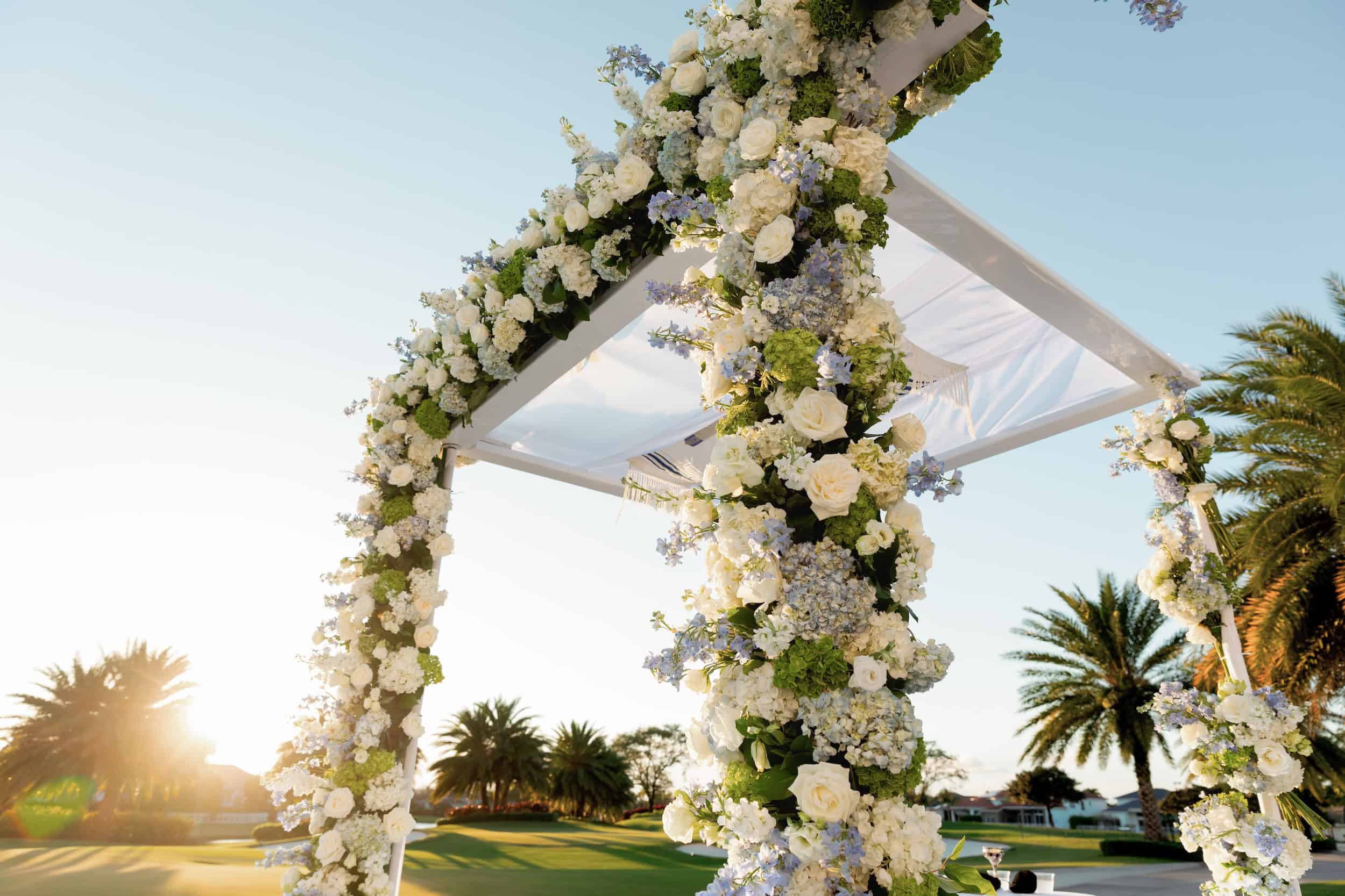 Elegant wedding arch decorated with white roses and greenery, set against a sunny outdoor backdrop with palm trees.