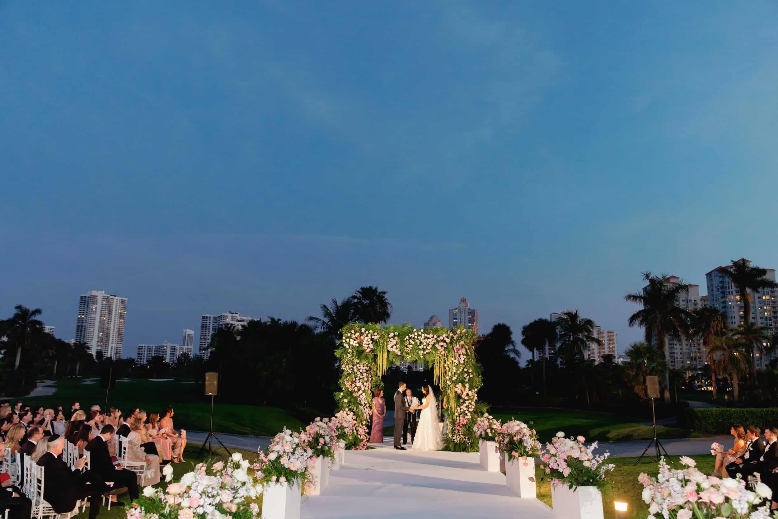 Bride and groom exchanging vows under a floral arch at an outdoor wedding ceremony.