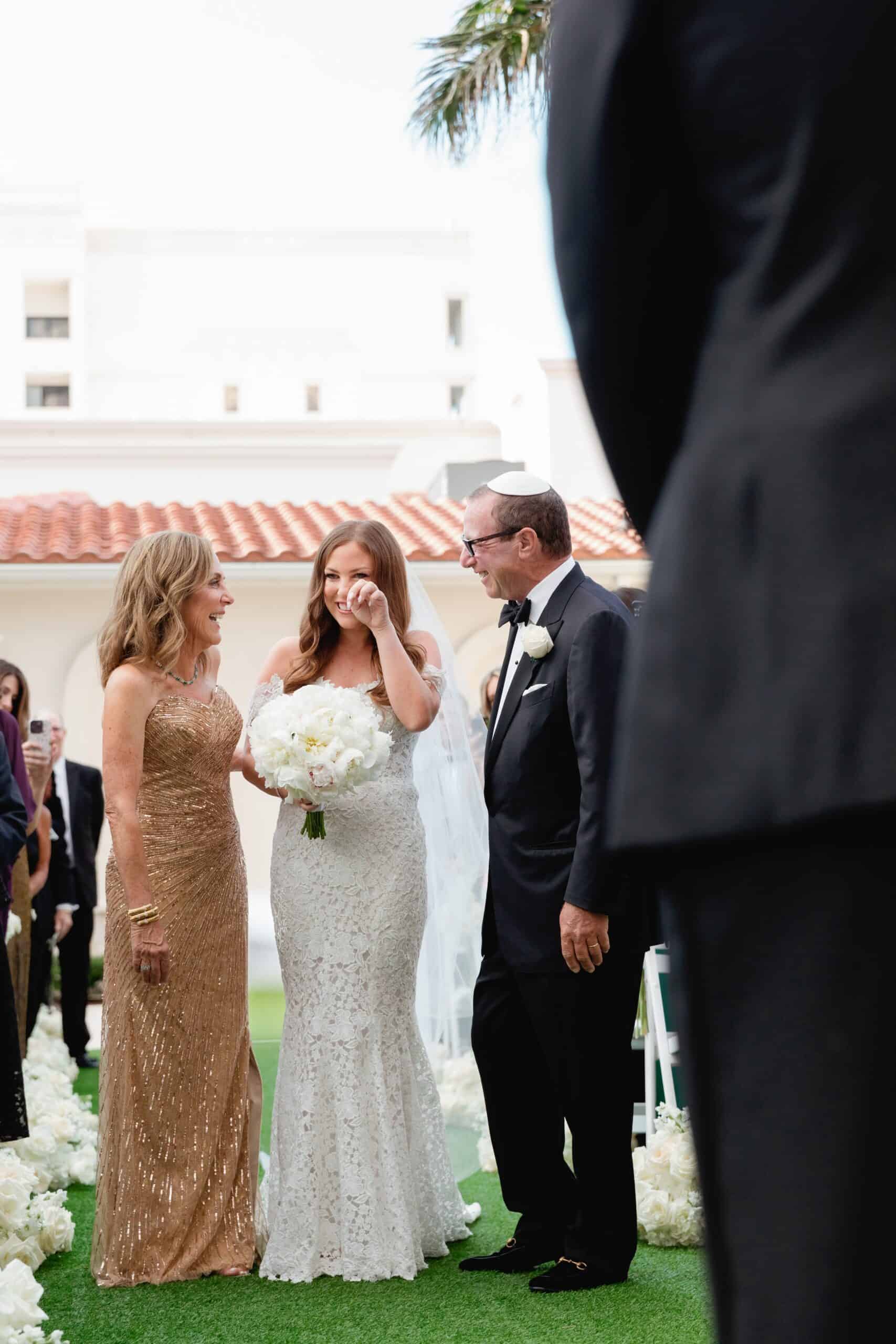 Bride in white gown and groom in tuxedo share a joyful moment with a woman in a gold dress at an outdoor wedding.