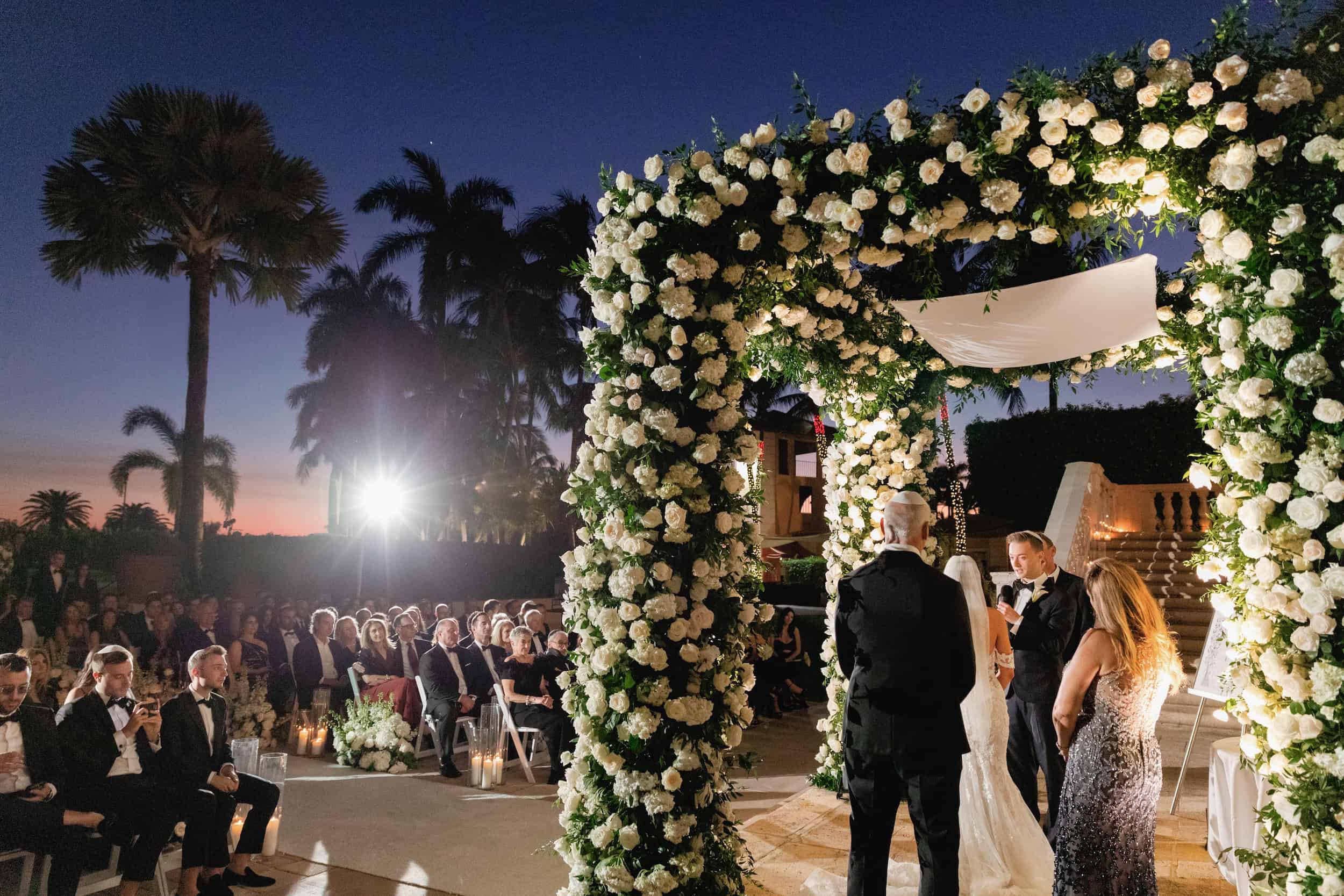 Bride and groom exchanging vows under a floral arch at sunset, guests seated nearby, tropical palm trees in the background.