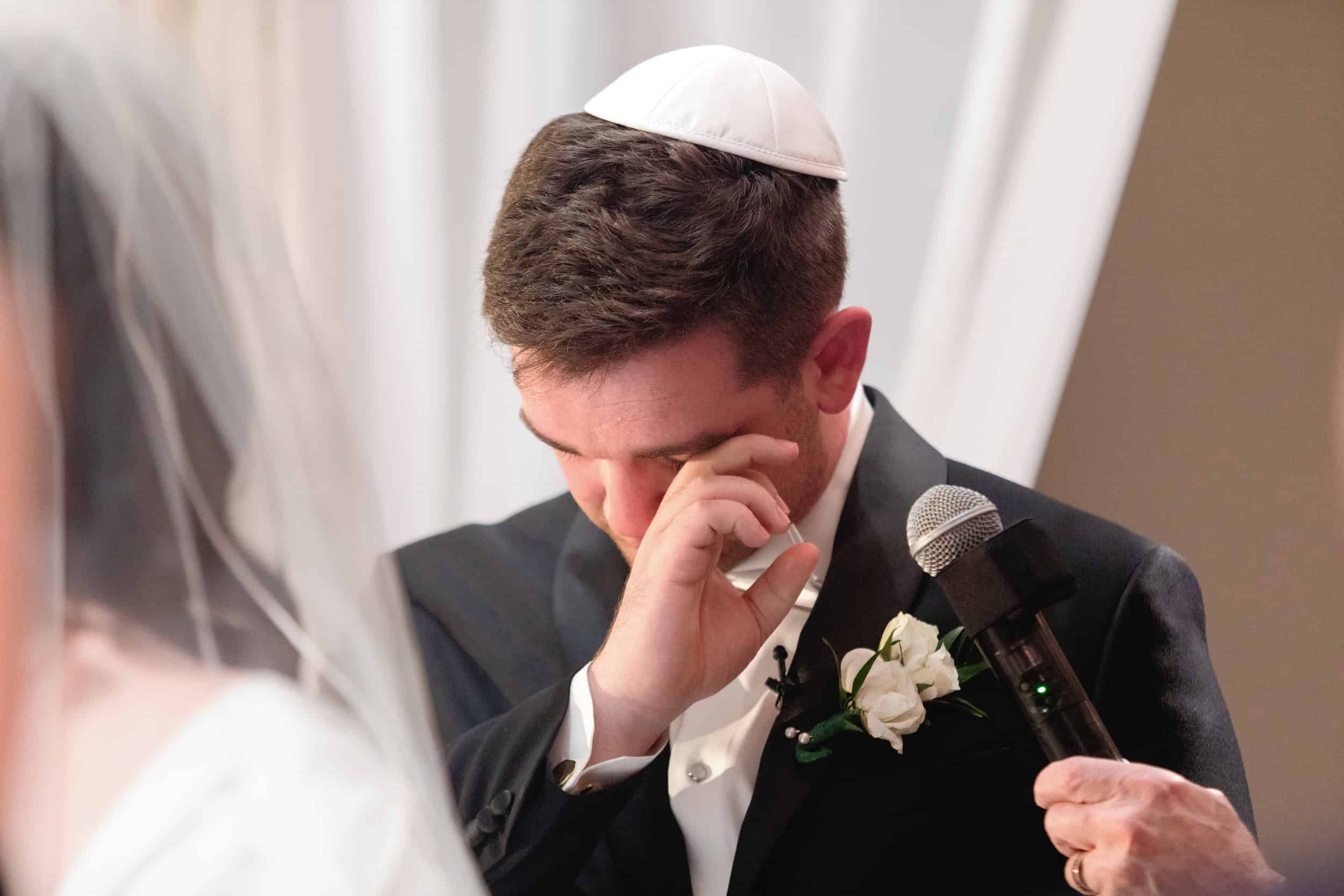 Groom wiping tears during emotional moment at wedding ceremony, holding a microphone, with white drapery in the background.