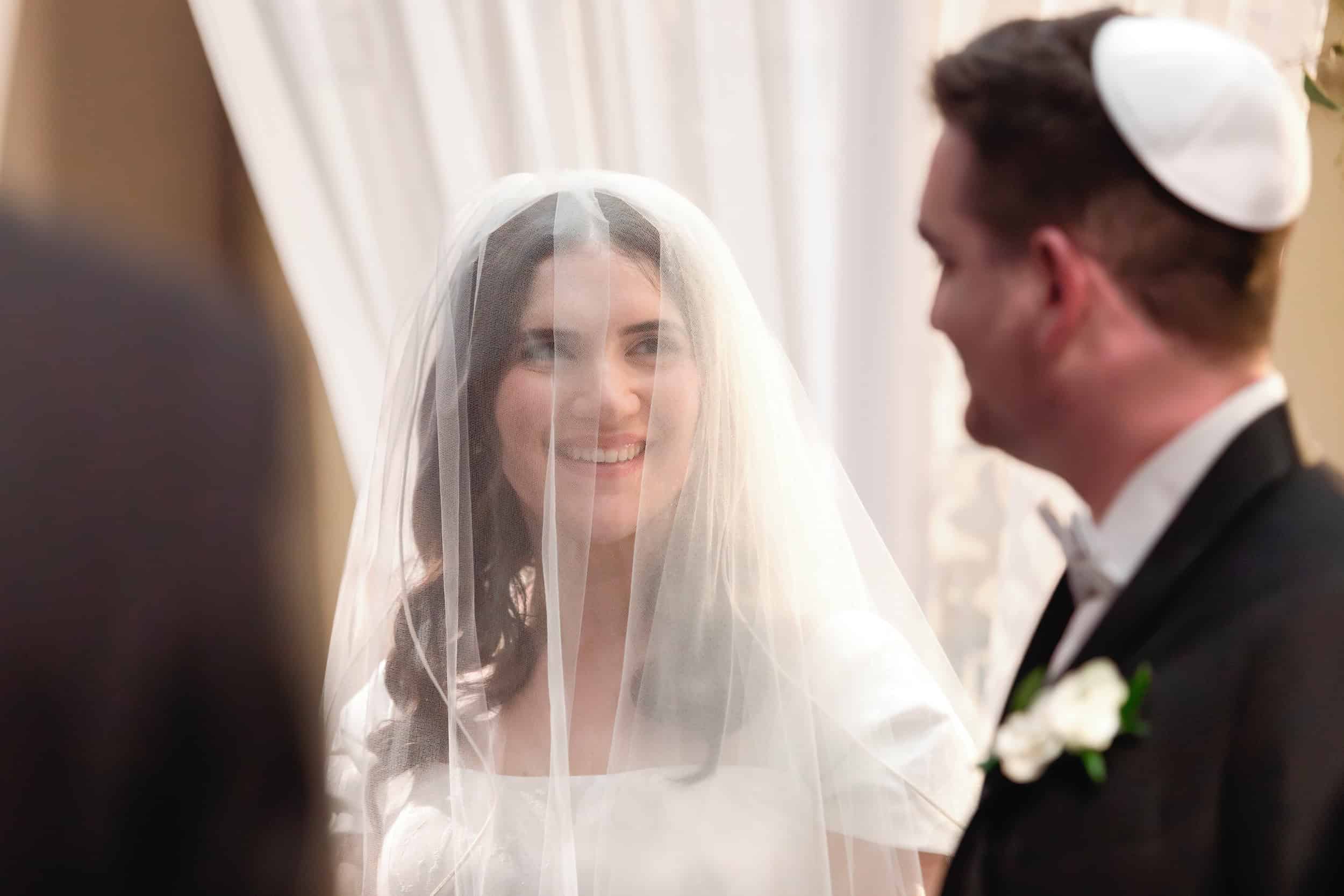 Bride with a veil smiling at the groom during their wedding ceremony, warm indoor lighting.
