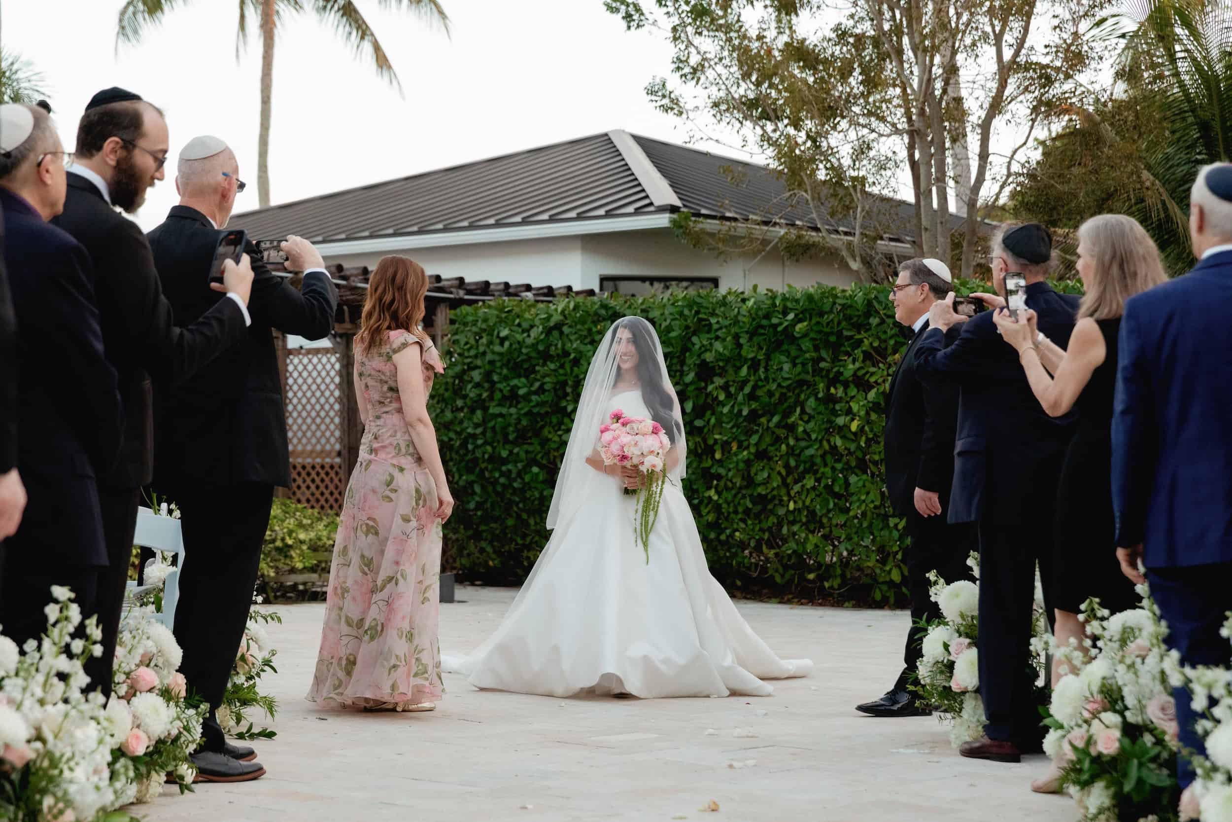Bride holding a bouquet of pink flowers, standing in front of guests at her wedding ceremony.