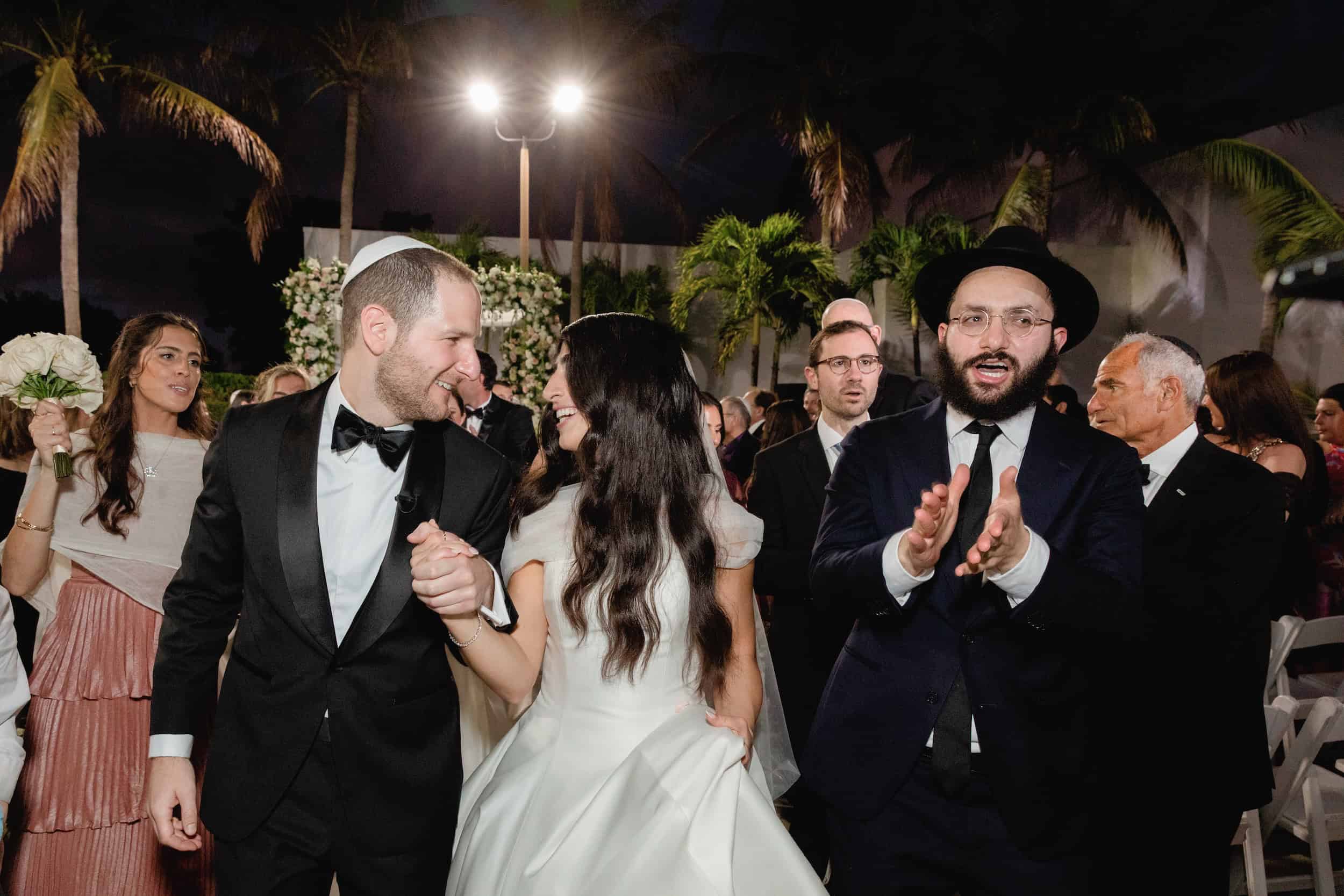 Bride and groom dancing with guests at a wedding reception, joyful expressions, nighttime outdoor setting with string lights.