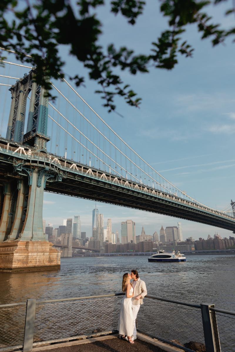 Engaged Couple Stands in Front of East River with Brooklyn Bridge and City Skyline in Backdrop